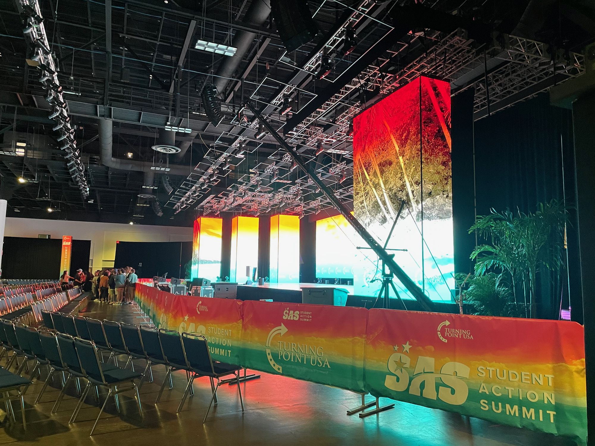 A camera rig rising on its boom from a base right of centre, in an exhibition hall lit blue and warm, with seating and drapery below.