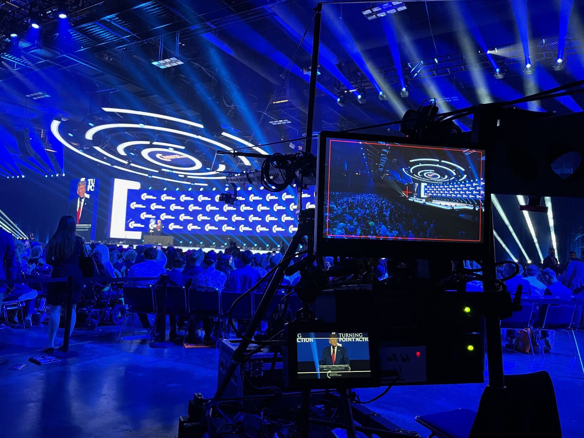 A broadcast camera on its head with an on-board monitor in the foreground, facing a blue-lit arena crowd and the stage beyond, during the 2022 Tampa run.