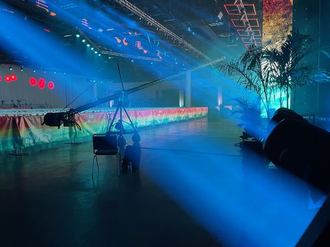 A camera on a Jib arm silhouetted in a darkened hall lit blue, colour panels on the screens behind.
