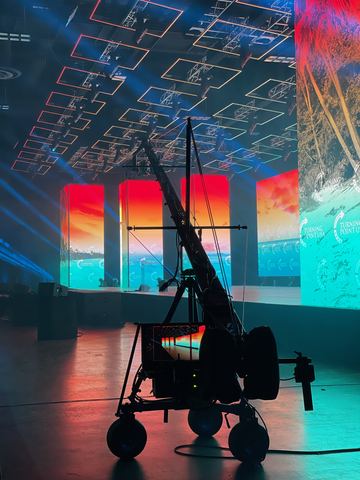 A camera rig on a wheeled base standing on a dark floor in front of a wall of red, blue and teal screens.