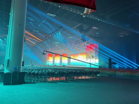 An empty arena of rows of seating facing a stage where a wide screen glows above sweeping blue light beams.