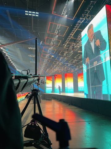 A camera on a tripod in the foreground of a dark hall, a large screen beyond it.