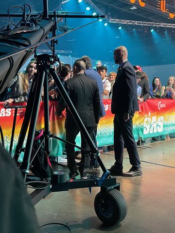 Crew standing beside a camera on a tripod on a stage, lit screens behind them.