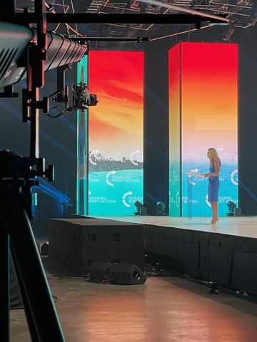 A camera on a tripod facing a stage with two tall screens.