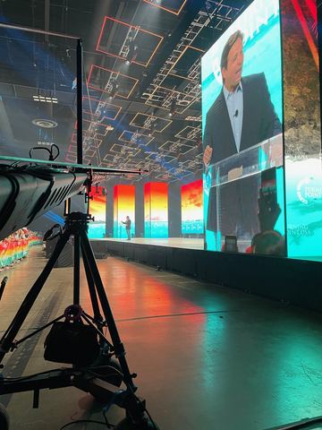 A monitor mounted on a tripod in front of a wide stage screen carrying red and blue graphics.