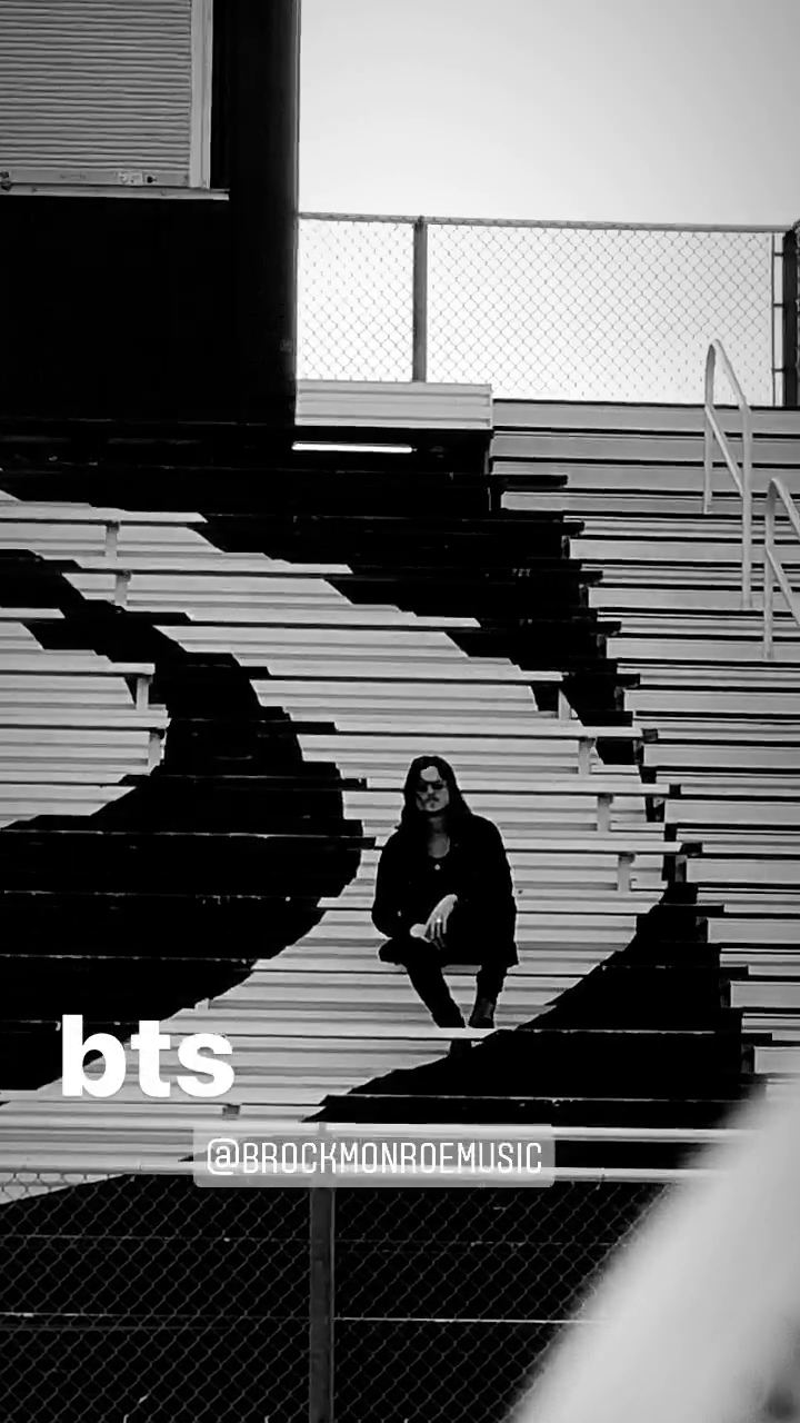 Brock Monroe, "Good Old Days" — behind the scenes. A black-and-white frame: a lone figure seated on stadium bleacher steps, chain-link fence and a building behind.
