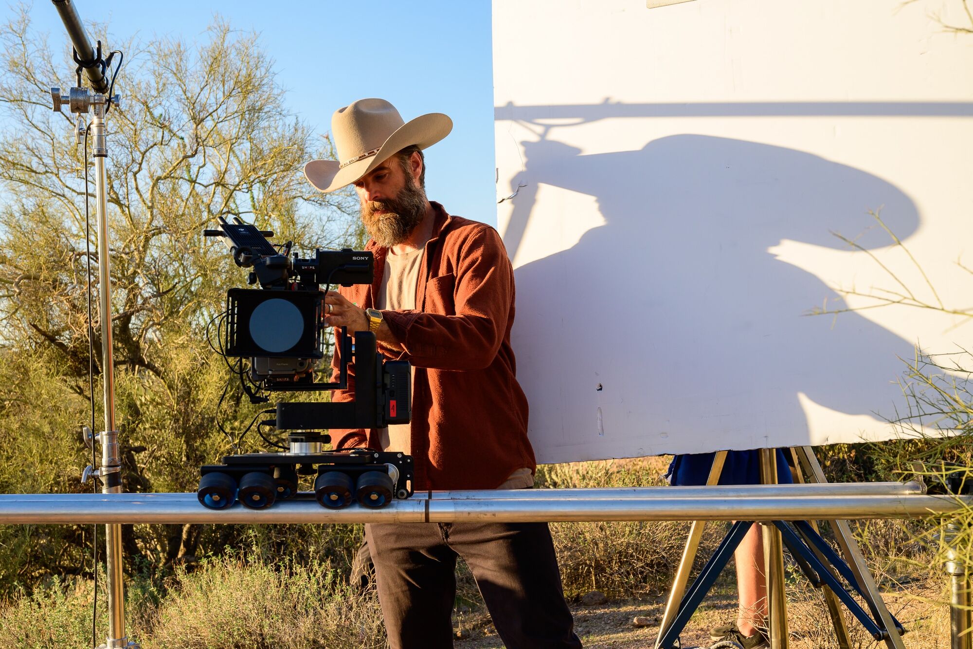 An operator at a cinema camera on a track-mounted dolly on a desert location in low golden-hour light, dry scrub and a bounce board behind.
