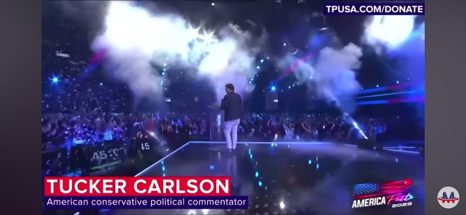 America Fest (December 2023) — Jib. Tucker Carlson on stage, with the broadcast's own graphics left in the picture: his name super, the America Fest mark and the event's donation URL.