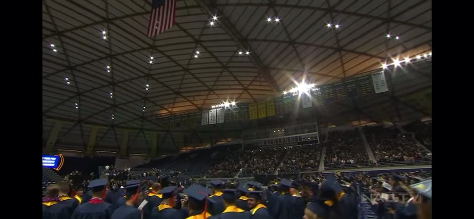 NAU Winter Commencement (2022) — Jib. From high in the stands: the graduates on the floor, the flag hanging from the roof structure and the tiers filled.